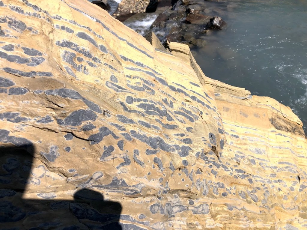There's some moving water visible in the background in the top right of the frame, but the image is dominated by a view of a rock formation. Yellow and orange bands of rock are punctuated by rows of dark gray stones. 