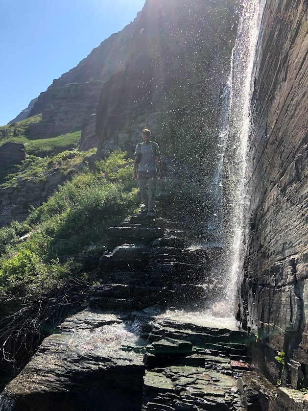 Dustin walks down a rocky ledge in the center of the image. To the right, a small waterfall sprays bright white droplets, backlit by the sun, onto the trail and into the air. 