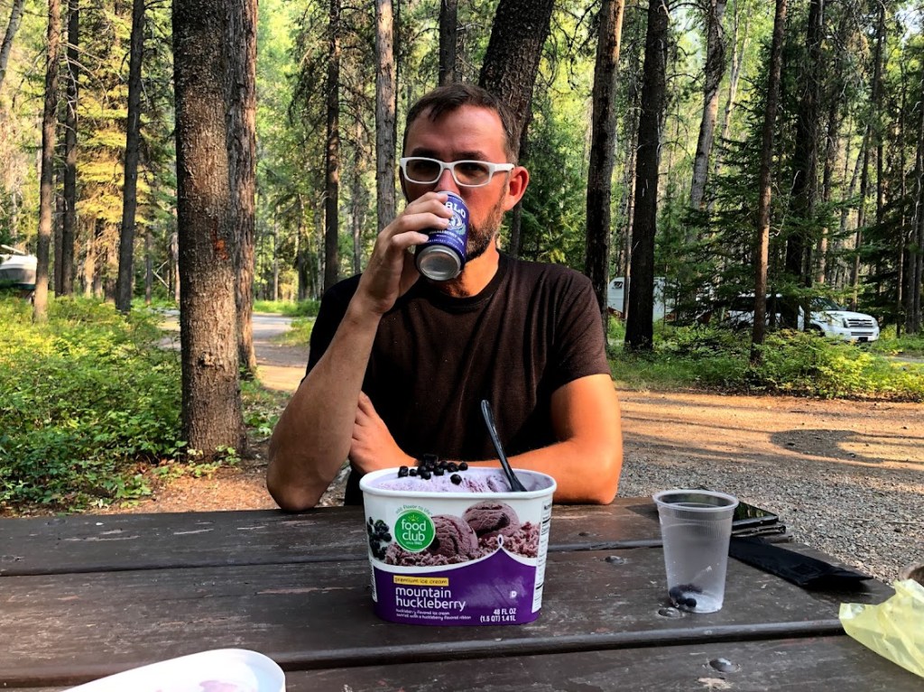 Dustin faces the camera in the center of the frame, sitting at a picnic table. He is drinking from a blue beer can. In front of him is a quart of Mountain Huckleberry ice cream, topped with fresh huckleberries. The background shows tree trucks and a few cars parked along the campground road.