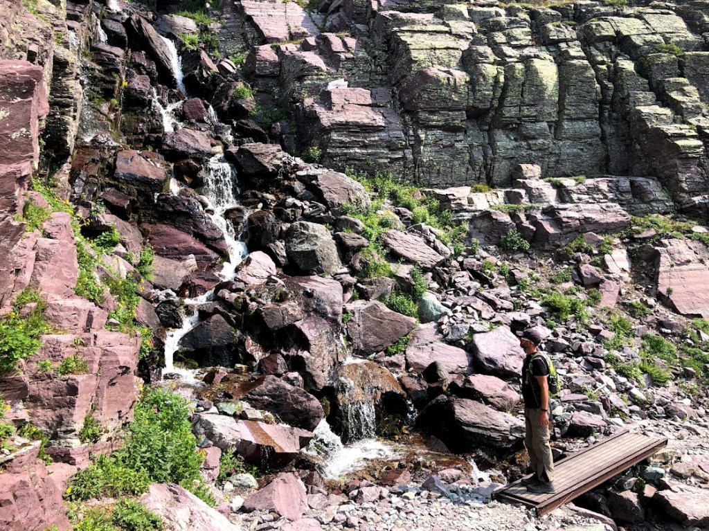 Dustin stands on a foot bridge over a waterfall stream and looks left and up, toward the waterfall as it comes down a rocky slope.
