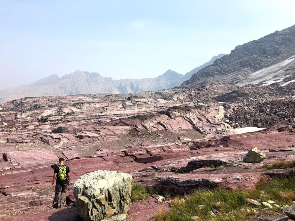 A red stone landscape nearly devoid of plants stretches in bands from side to side. Dustin stands bottom left, his back to the camera.