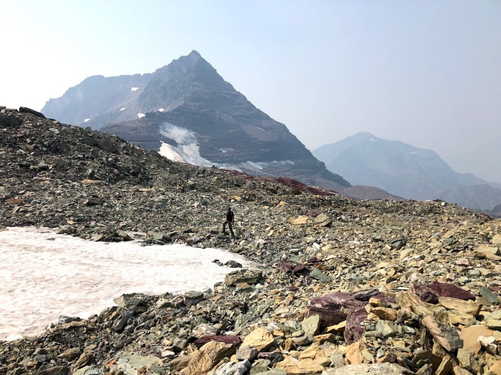 Dustin, very small, stands at a distance in a field of glacial till, walking away from the camera and from Sperry Glacier.