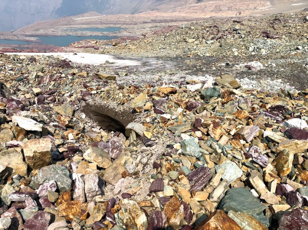 Colorful rock rubble fills the frame with several small glacial pools in the far background. At the center of the photo, an ice bridge can barely be discerned under the rocks. 