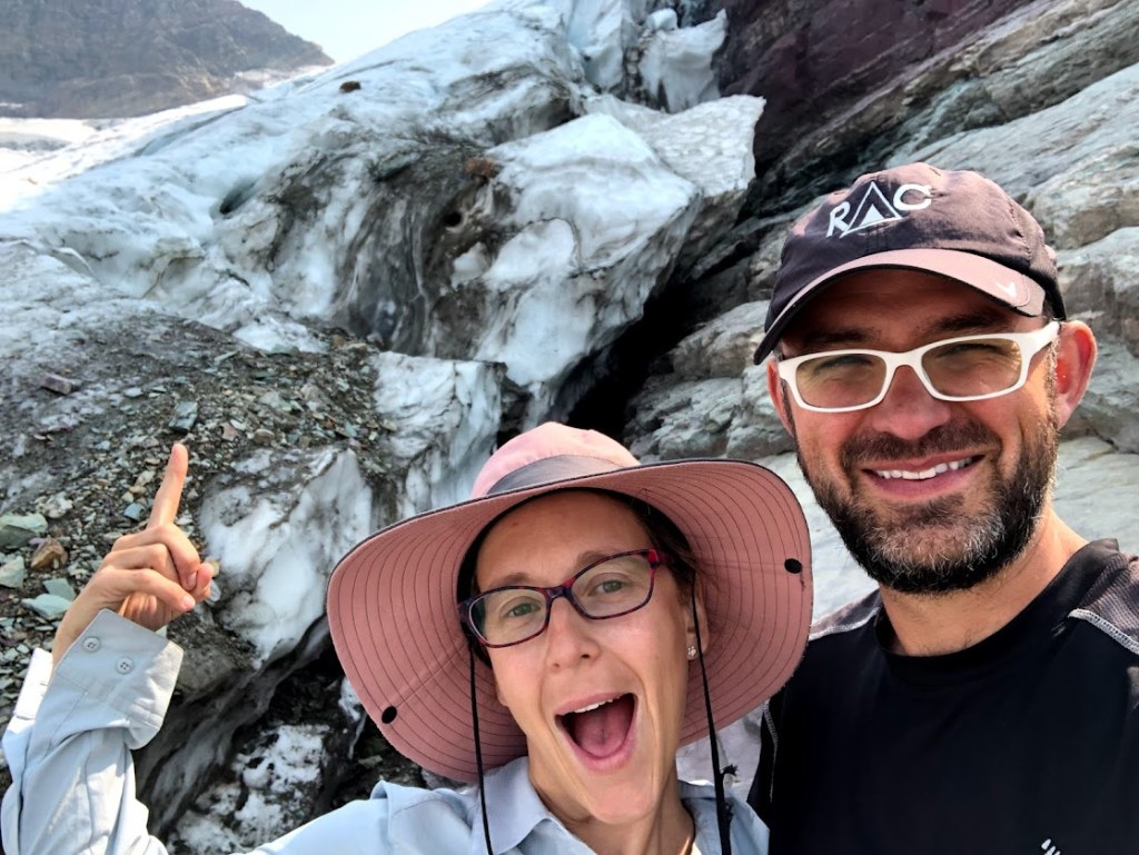 Selfie of Dustin and Laura in front of the glacier. Laura is wearing a goofy grin and pointing at the glacier behind her. Dustin is laughing at Laura. 