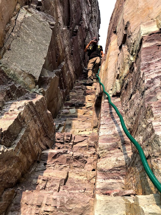 A narrow gap between two vertical stone cliffs contains stairs that lead through Comeau Pass. Dustin stands on the stairs, facing the camera at center, but watches his feet instead of the camera because he believes in safety. 