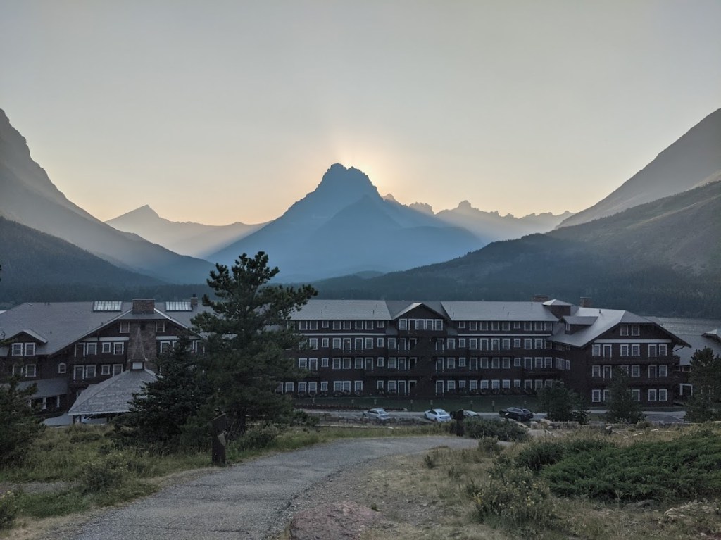 The four-story dark brown outline of the Many Glacier hotel fills the bottom third of the image, stretching off either side of the frame. Above it, there are dark silhouettes of mountains sloping away from the building, backlit by the sun that's just set behind a peak at the very middle of the image. 