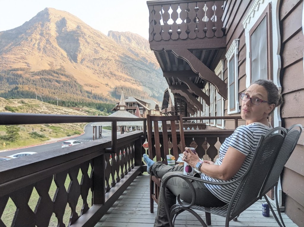 Laura sits in a chair on a dark brown porch on the right side of the frame, looking at the camera and holding a can with a stylized huckleberry on it. A mountain rises above her in the background on the left side of the image. On the right, the side of the hotel stretches away into the distance, with other balconies and porches visible hanging off the side of the building. 