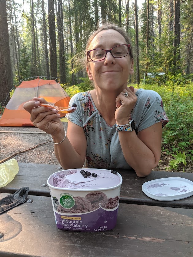Laura sits at a picnic table, smiling at the camera. Her elbows are on the table, one hand holding a spoon full of huckleberry ice cream with one plump, fresh berry sitting on top. The quart of ice cream sits on the table in front of her. In the background, the orange tent sits to the left, framed by evergreen tree trunks. 