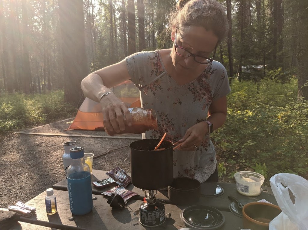 Laura stands in front of a picnic table at center, facing the camera but looking down at the pasta sauce she is pouring into the pot on the camp stove. Sun comes through tree trunks behind her. 