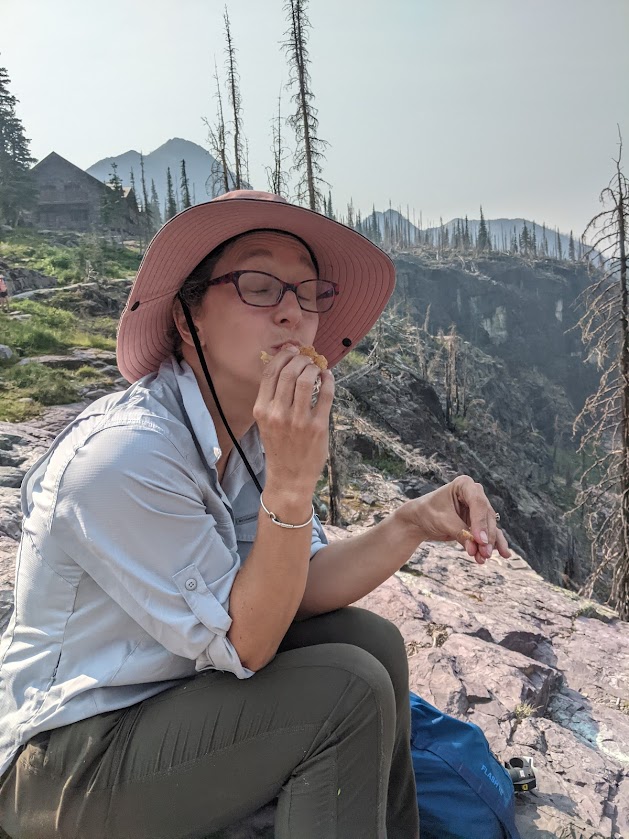 Laura sits in the center of the frame, wearing a hat, eating a sandwich, eyes closed in contentment. A cliff drops off behind her, topped with burned pine trees. 