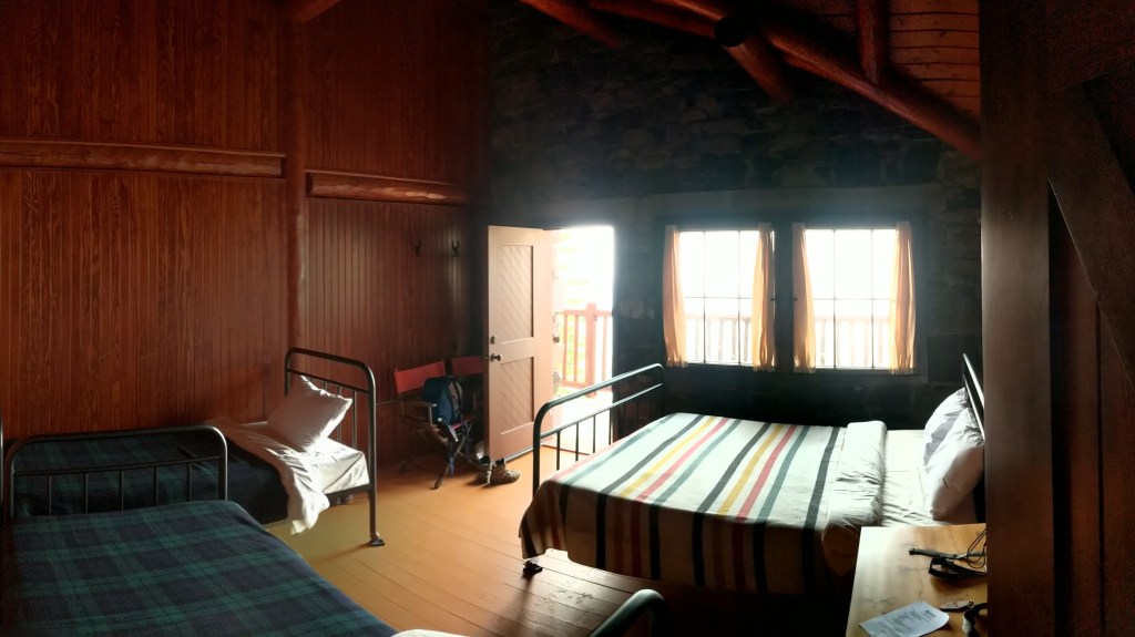 A Sperry Chalet dorm room with a double bed on the right and two single beds on the left. The shot looks out the windows to the porch beyond. The light coming through the windows makes it difficult to see details. 
