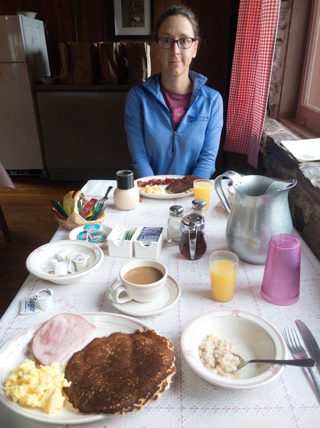 Laura sits opposite the camera at a table in the Sperry Chalet dining hall, looking at the camera with a bemused smile. The table is set with a large breakfast.