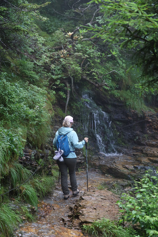 Laura hikes away from the camera, in the center of the frame, along a wet rocky path. A series of streams cascades down the rocks that border the path to her left. 