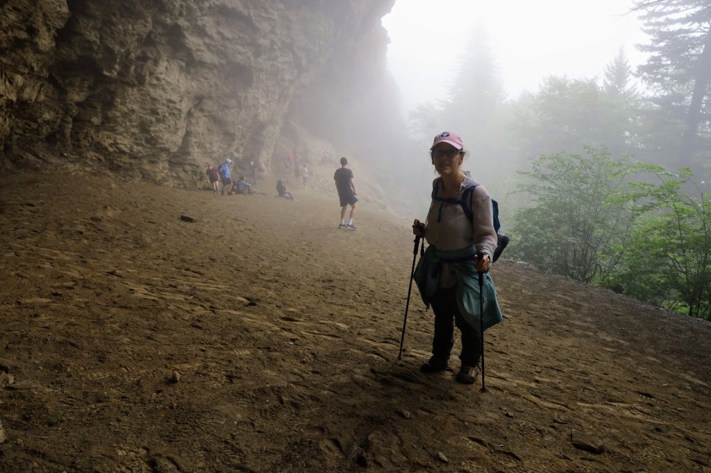 Laura stands on a sandy slope in the center-right of the frame, facing the camera. In the background, a cliff rises up, leaning outward to form a kind of overhang. Smaller people can be seen along the bottom of the cliff in the background. 