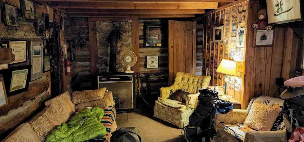A room lit only by lame fills the frame, with a worn couch on the left and a couple chairs covered with mismatched pillows on the right. The plank walls are covered by certificates and photos.