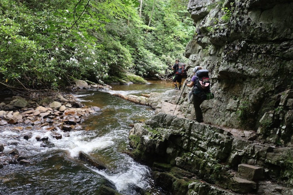 The creek runs around a rocky cliff-face which fills the right half of the frame. Hobz, followed by Laura, walk along a narrow path between the water and the cliff. 