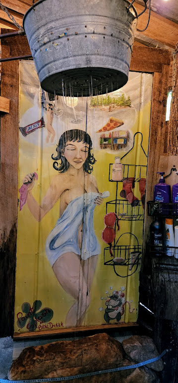 A shower stall with the back wall painted with: a mostly naked lady holding a towel loosely around herself. She is dreaming of a snickers bar, a naked man, a slice of pizza, a glass of wine, a stand of pine trees, and a cartoon rat covered in soap bubbles. A tin bucket hangs at the top of the frame, water pouring out of the many holes punched in it because this, as it turns out, is the shower head.