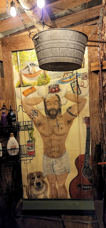 A shower stall featuring a back panel painted with a mostly naked man washing his hair, surrounded by a dog, a guitar, a beer, a canoe, a hammock, candy, and more beer. A tin bucket full of holes is at the top of the frame.