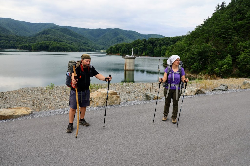Hobz and Laura stand on the dam road with the reservoir in the background. They are both looking pensively off to the right.