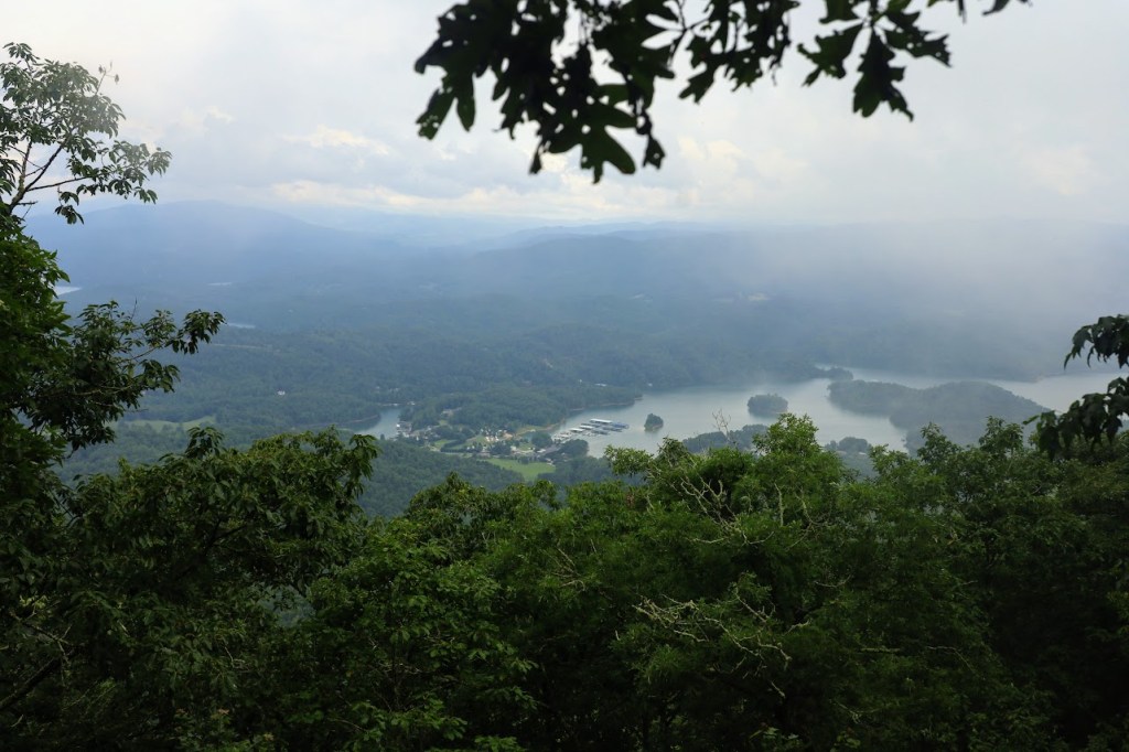 A high view down into the valley with part of the reservoir visible. Boats and buildings can be seen on the reservoirs left side. The sky is very overcast.