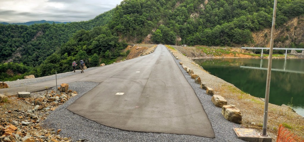 An asphalt road stretches across a dam from the bottom of the frame toward the back. The water of the reservoir is to the right, the drop-off to the left. Hobz and Laura, very small, can be seen toward the middle of the road walking away from the camera.