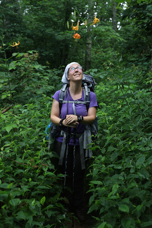 Laura stands center, surrounded by green undergrowth, looking up at a flower that grows from a very tall stalk and dangles face-down, as if looking back at her. 