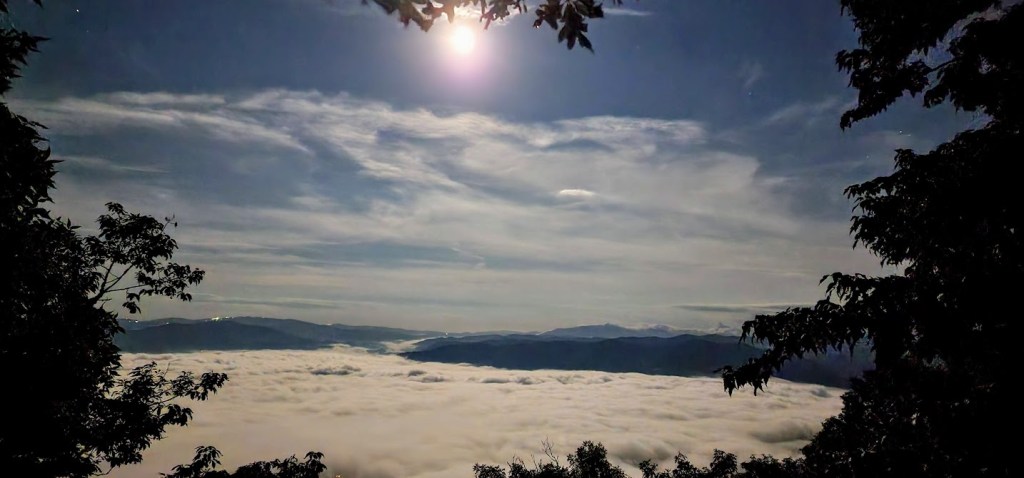 A photo showing the same reservoir valley that was shown above. The entire valley is filled with fog, with hills rimming it in the background. A nearly full moon shines down from the center top of the photo, lighting up the fog.