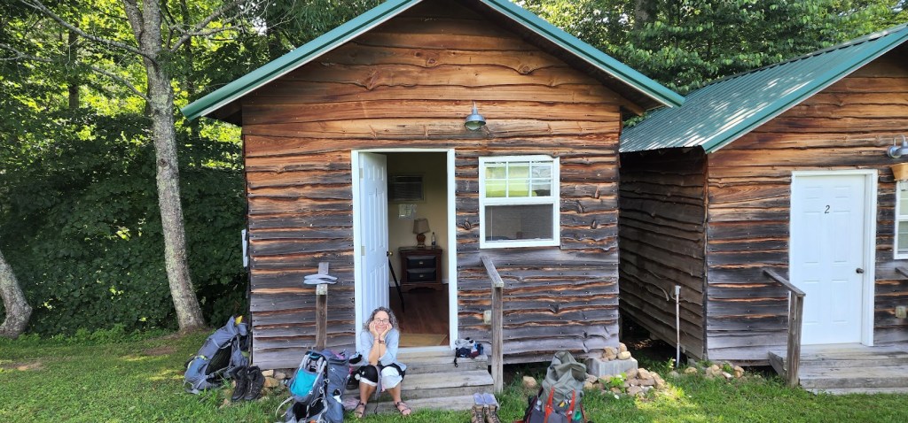 A photo of the front of one of the small cabins with Laura sitting on its front steps, chin in her hand, smiling at the camera. The door to the cabin is open, and a small dresser with a lamp can be seen inside. Backpacks and gear are scattered on the grass in front.