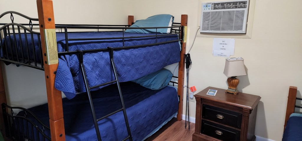 A pair of bunk beds against the cabin wall to the left, a small dresser with a lamp center, and the edge of a third bed barely visible to the right. The walls are very white, and an AC unit sits above the lamp. 