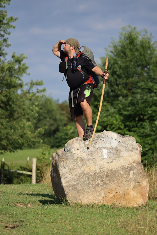 Hobz poses on top of a boulder, walking stick in one hand, other hand held up to shield his eyes as he looks toward the left of the frame.