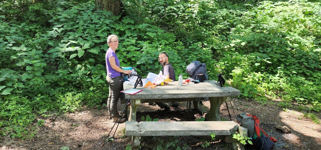 A stone picnic table sits at center. Dustin sits on the bench farther from the camera while Laura stands on the left end of the table, sorting through packages of food for lunch. Both are looking at the camera. 