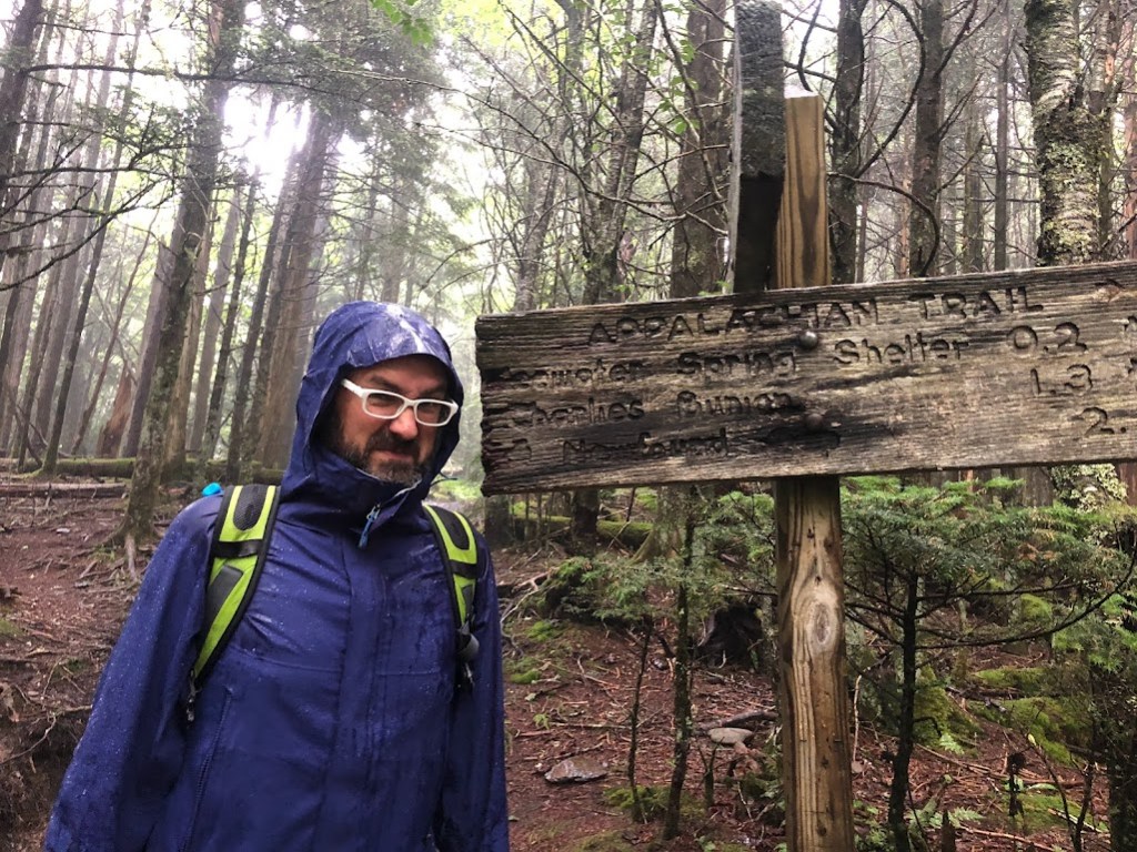Dustin, in a very wet rain jacket, stands next to a trail sign that says:

APPALACHIAN TRAIL
Icewater Spring Shelter 0.2
Charlies Bunion 1.3
Newfound Gap 2.7