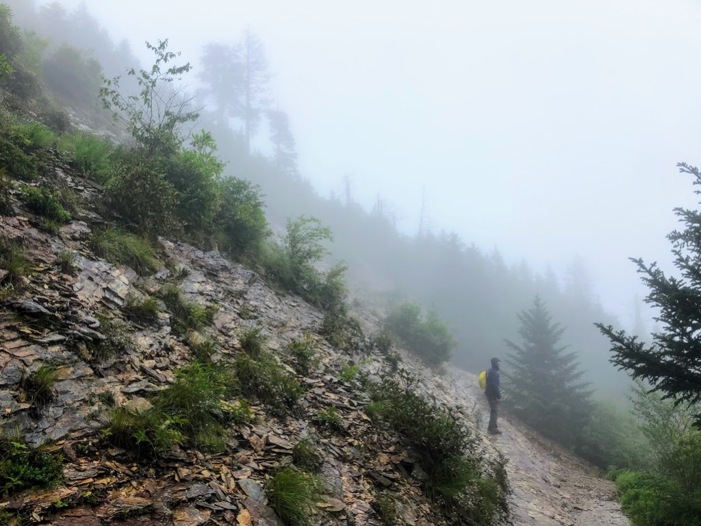 Dustin stands on the side of a rocky slope looking out at a view hidden by fog. 