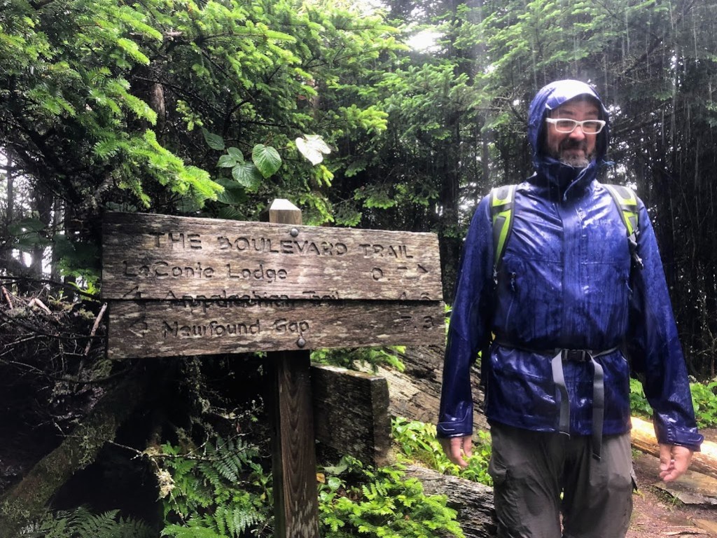 Dustin stands to the right of a sign that reads:

THE BOULEVARD TRAIL 
Le Conte Lodge  0.7 [right arrow]
[left arrow] Appalachian Trail 4.6
[left arrow] Newfound Gap 7.3

Rain is pouring down so heavily it appears in the photo as vertical white stripes. Dustin is soaked.