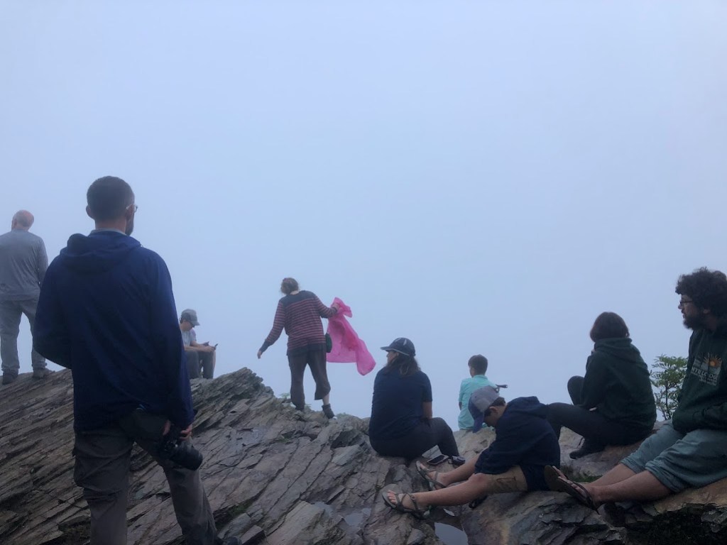A crowd of people sit and stand on a rocky ledge. The view over the ledge is only fog.