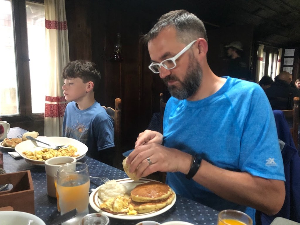 On the right side of the frame, Dustin sits at a table in front of a plate full of pancakes, scrambled eggs, and grits. He is cutting open a biscuit. A young boy sits to Dustin's right, on the photo's left, also in front of a large plate of food. 