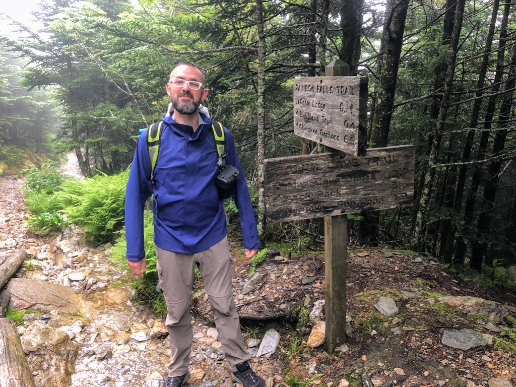 Dustin stands to the left of a pair of trail signs that can't quite be read because they are wet and dark. A rocky trail slopes downward toward the back of the frame to Dustin's left. Dustin looks happy.
