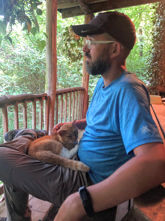 Dustin sits on a bench facing left over a wooden porch rail. He pets an orange cat that is curled up in his lap, looking content.