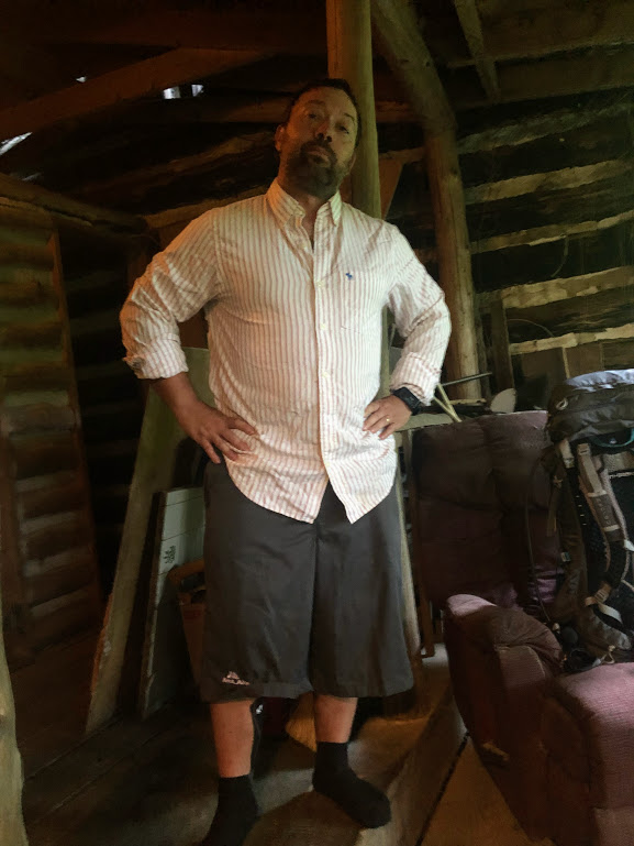 Hobz stands in the center of the frame. He is wearing a striped white shirt and a pair of shorts that come down to the middle of his calves. The background is obscured by lack of light.