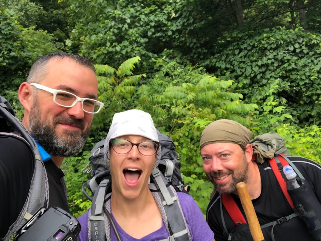 A selfie against a lush, green, background. Dustin is to the left, Laura center, Hobz right. All are wearing backpacks. Hobz and Laura wear bandanas. Everyone looks happy. 