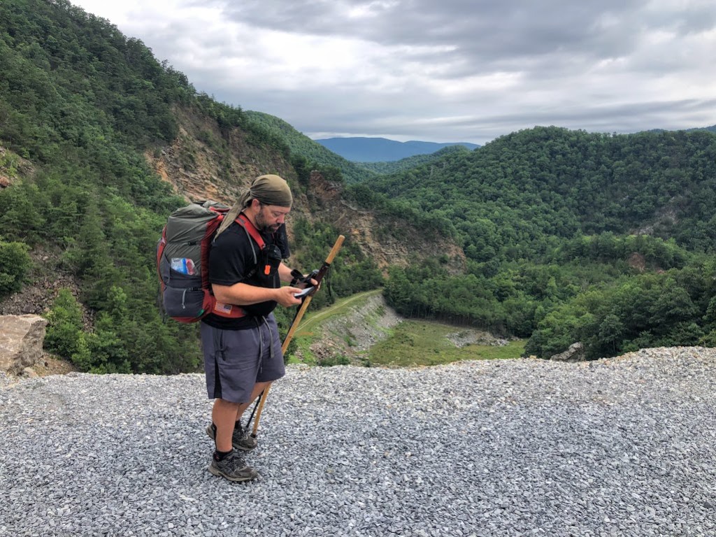 Hobz stands on the edge of the dam road with the drop-off and valley behind him. The sky is overcast. Hobz is consulting his phone.