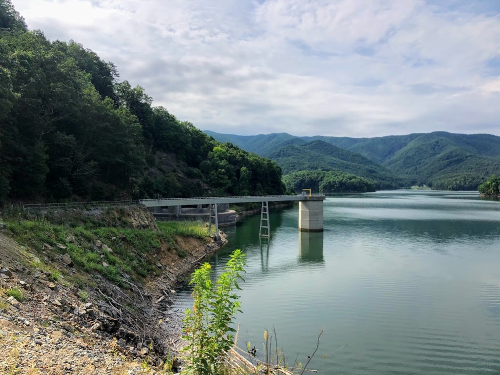 A photo of the reservoir, showing the bank to the left with a metal pier stretching out in the middle distance. The water is gray, and the hills behind it covered in dark green trees.