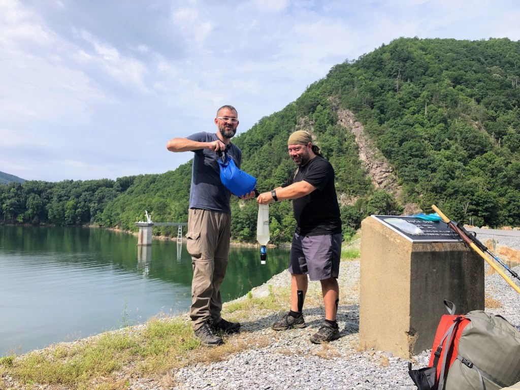 Hobz and Dustin, standing on the edge of the reservoir. Hobz holds a water filtering back at waist height while Dustin using a water-collecting bag to fill it. Both look at the camera because apparently Laura said something to get their attention.