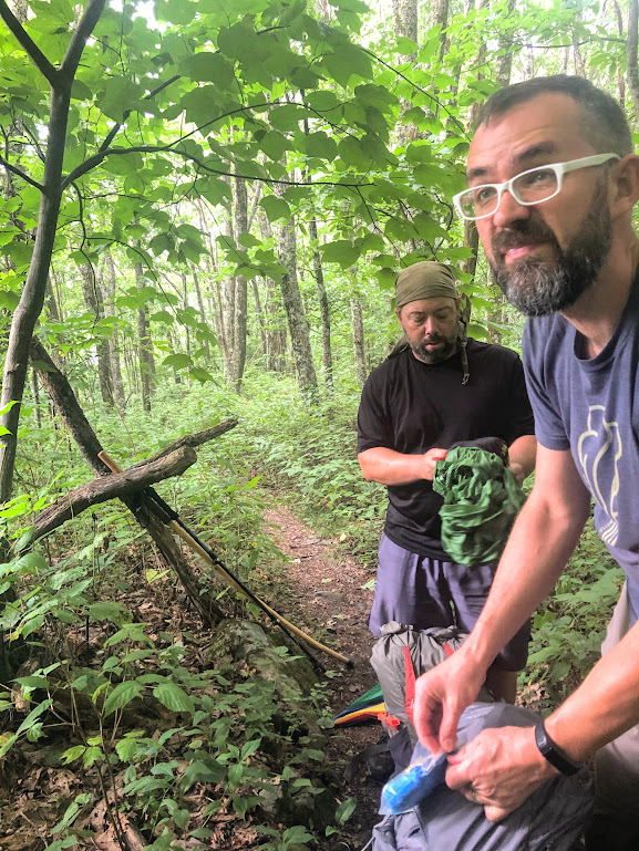 Standing under bright green-leafed branches, Hobz and Dustin pull their rain gear out of their backpacks. Dustin is peering speculatively toward the left.
