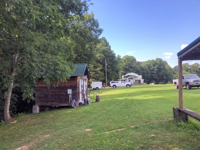 A grassy field stretches out under a blue sky, with small log cabins to the left and the post of some large building to the right. Several trucks are parked in the background.