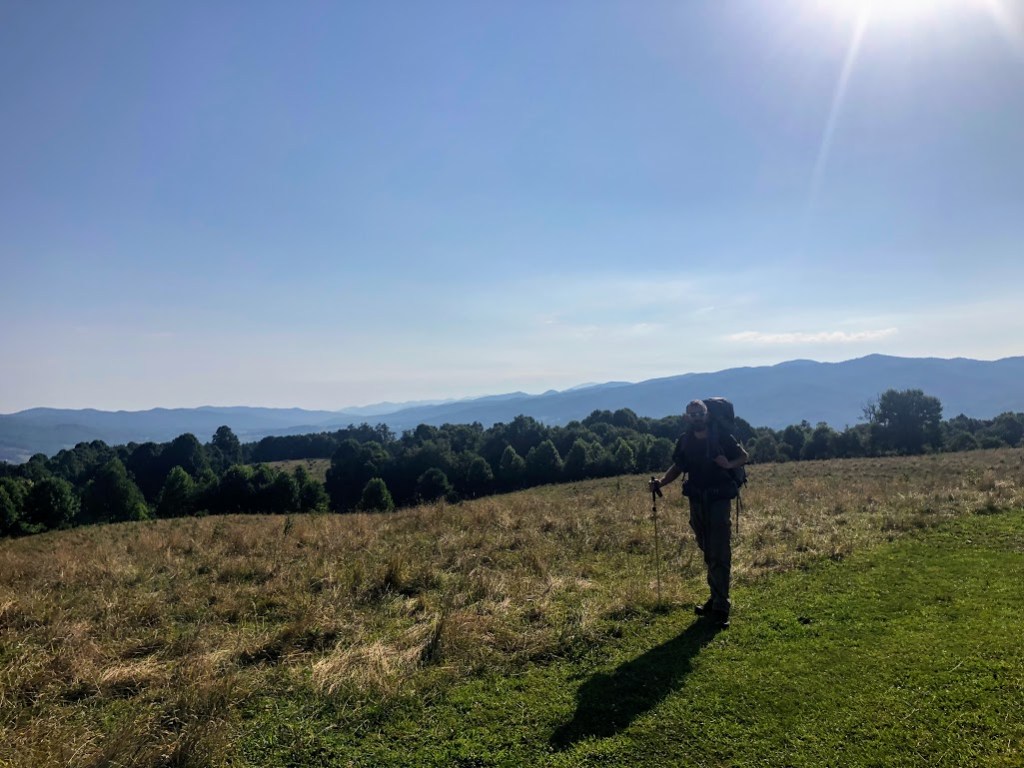 Dustin stands on the edge of a grassy field, bottom right of the frame, with rows of mountains becoming more faded with distance in the background. The sun is visible at the top right of frame, lighting the field and leaving Dustin in shadow. 