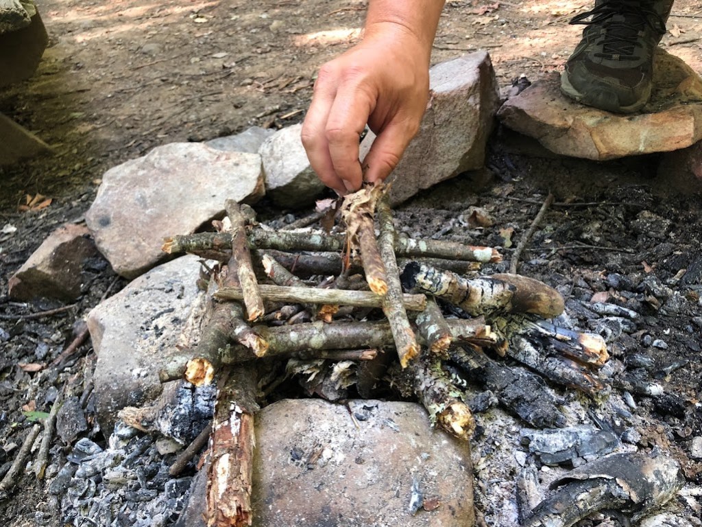 Close up of a stone-ringed fire circle. A stack of broken sticks has been set up in the shape of a hashmark several layers tall. A hand belonging to Hobz is adding another stick to the pile. 