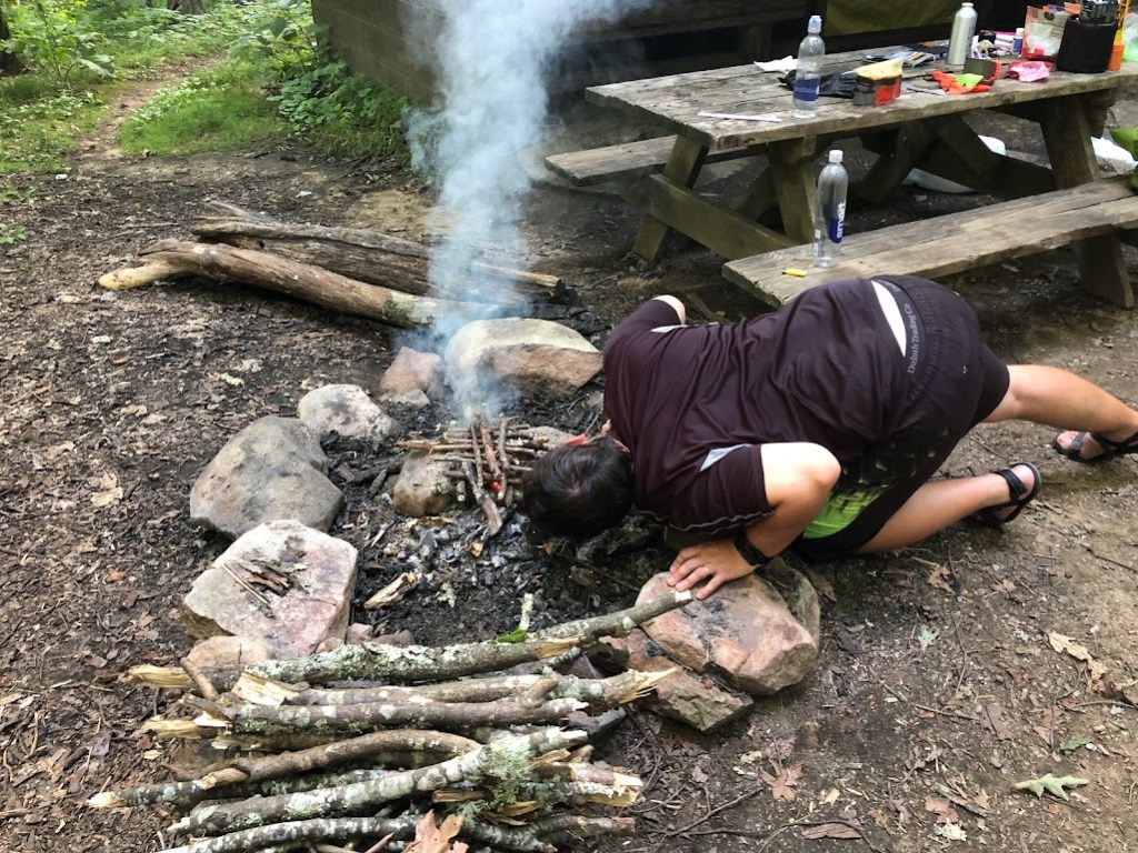 Hobz leans over the stone-ringed fire circle, body turned away from the camera, head close to the pile of sticks. Smoke rises from the sticks and a very small bit of orange flame can be seen between them. A picnic table covered in water bottles and cooking supplies is in the background. 