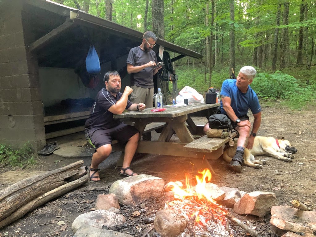 Hobz sits on the left side of the frame at the picnic table, Dustin standing to his right, and a white-haired stranger named Hashtag sits opposite Hobz, on the right of the frame. A yellow dog lays at Hashtag's feet. The fire circle with the fire burning inside it is in front of the picnic table, at the bottom of the frame. The shelter is in the background. 