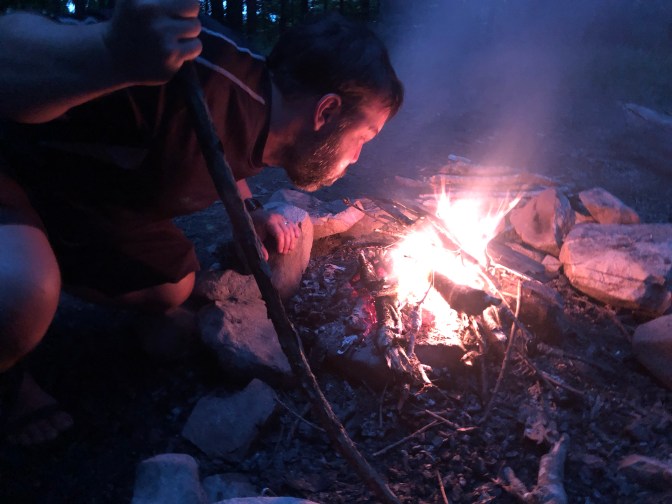 Night time photo of Hobz, on the left of the frame, leaning over the fire and blowing. 